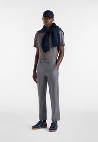 Muž oblečený v šedém tričku, šedých kalhotách, námořnických modrých teniskách, námořnické mikině přehozené přes ramena a námořnické kšiltovce na bílém pozadí.