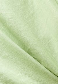 Tissu en lin vert clair doux avec texture de tissage visible et plis délicats formant des lignes diagonales.