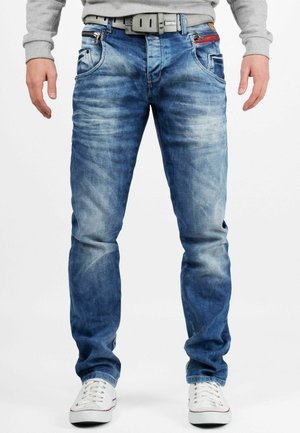 Lichte blauwe denim jeans met een vervaagd uiterlijk. Kenmerken zijn onder andere vijf zakken, een ritszak en een grijze riem. Draagt met witte schoenen.