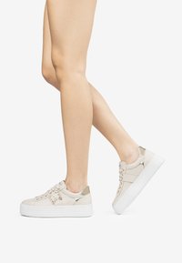 Beige sneakers med en tekstureret overflade, som har en platformssål, metaldetaljer på hælen og snørebånd med dekorativ hardware.