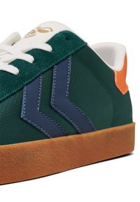 Grüner Textilsneaker mit weißen Schnürsenkeln, marineblauen Akzentstreifen, orangefarbener Fersendetail und einer Gummisohle. Strukturierte Oberflächen.