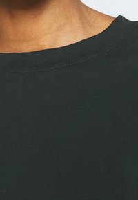 Gros plan sur le cou et l'épaule d'une personne portant un t-shirt vert foncé à col ras du cou avec des coutures visibles.