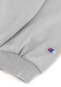 Grijze sweatshirtstof met ribbelmanchetten. Bevat een klein, geborduurd logo in blauw, rood en wit. Zachte textuur, casual ontwerp.