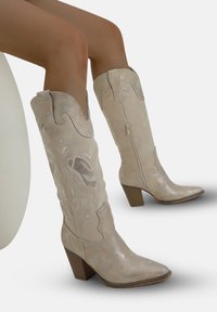 Stivali da cowboy color beige al ginocchio con finitura lucida, cuciture decorative, punta affusolata e tacco spesso. Cerniera laterale per una facile calzata.