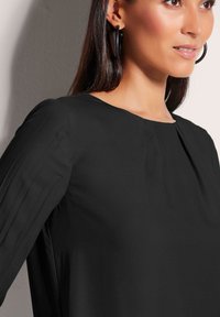 Schwarze Bluse mit langen, gefältelten Ärmeln und rundem Halsausschnitt. Glatte Textur mit minimalen Details und einer lockeren Passform.