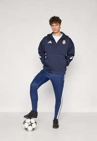Marineblaues Zip-Jacke mit weißen Akzenten und Real Madrid Logo, kombiniert mit passenden Jogginghosen mit weißen Seitenstreifen und schwarzen Fußballschuhen, die auf einem Fußball stehen.