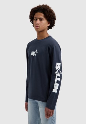 Jonge man met krullend haar die een marineblauw shirt met lange mouwen draagt met een wit "B*LN"-logo op de borst en de linker mouw, gecombineerd met lichtblauwe spijkerbroek.