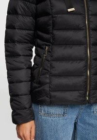 Giacca a vento nera con texture trapuntata, zip dorate e tasche laterali. Presenta un colletto alto e un design elegante e aderente.