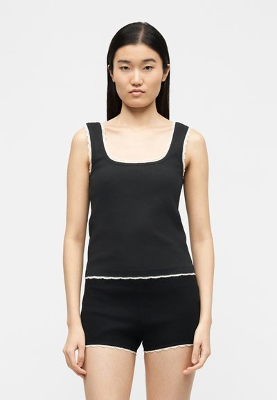 Jeune femme aux cheveux lisses et foncés portant un haut noir sans manches et un short noir, tous deux ornés d'une bordure en dentelle blanche, se tenant devant un fond uni.