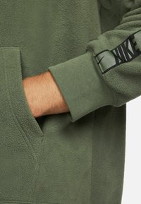 Grüner Fleece-Pullover mit Kängurutasche, gerippten Bündchen und einem schwarzen Nike-Logo auf dem Ärmel gedruckt. Weicher Stoff, lässiges Design.