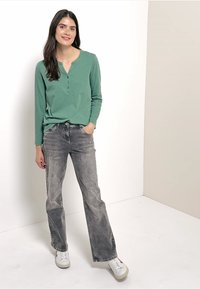 Vihreä pitkähihaisena henley-paita nappien kera, yhdistettynä haalistuneisiin harmaisiin leveälahkeisiin farkkuihin ja valkoisiin tennareihin, tasaisen taustan edessä.