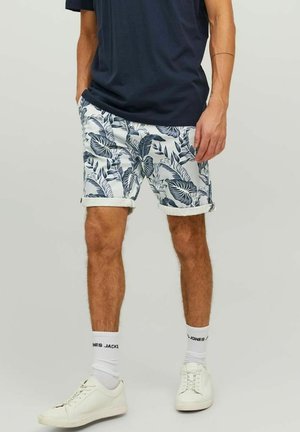 Heren korte broek met een witte basis en een blauw tropisch bladerenpatroon, opgerolde mouwen, gecombineerd met een marineblauw shirt en witte sneakers.