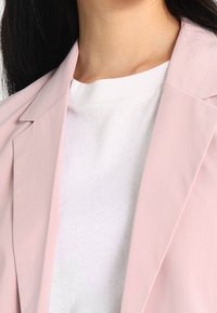 Světle růžový blejzr vyrobený z hladké tkaniny, s ostrými klopami a jednoduchou siluetou, přehozený přes bílou košili s kulatým výstřihem.