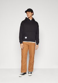 Black hoodie with a kangaroo pocket, paired with orange-brown pants and black sneakers. Simple design, cotton material, relaxed fit.