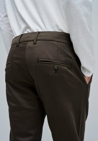 Brązowe spodnie o gładkiej fakturze, z dwiema tylnymi kieszeniami z zapięciem na guziki oraz eleganckim fasonie. Połączone z dopasowaną białą koszulą z długim rękawem.