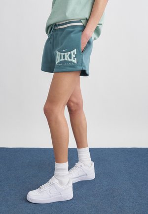 Persoon draagt turquoise Nike sportshorts, witte sokken en witte Nike sneakers, staand op een blauw tapijt met hand in broekzak.