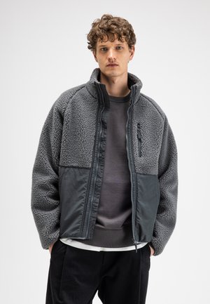 Young man wearing a gray fleece jacket with zipper and pockets, standing against a plain white background.