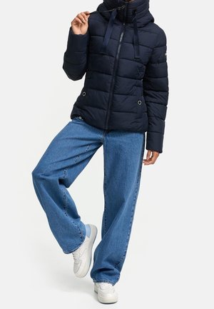 Personne portant une doudoune bleu marine avec cordons, un jean bleu ample et des baskets blanches, levant légèrement une jambe vers l'avant.