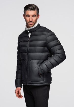 Mann trägt schwarze gesteppte Steppjacke mit Reißverschlusstaschen, steht mit einer Hand in der Tasche und schaut nach vorne auf neutralem Hintergrund.