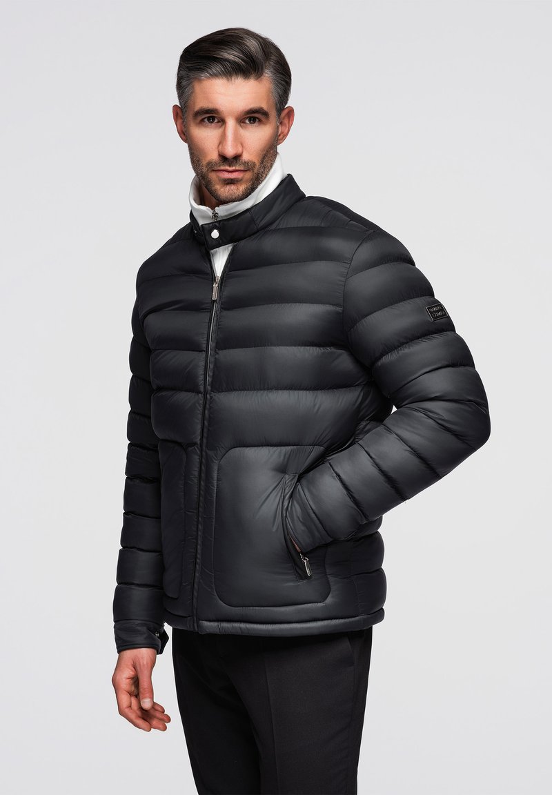 Homme portant une veste matelassée noire avec des poches zippées, debout avec une main dans la poche et regardant devant sur un fond neutre.