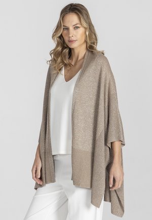 Kobieta o falowanych blond włosach, ubrana w jasnobeżowy, otwarty kardigan na biały top z dekoltem w serek oraz białe spodnie, stojąca na tle jednolitego szarego tła.