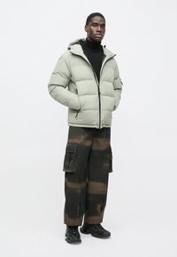 Vaaleanvihreä puhallettava takki hupulla, mustalla vetoketjulla ja sivutaskuilla. Yhdistetty mustiin cargo-housuihin, joissa on ruskea sidontakuosi, sekä mustiin kenkiin.