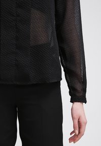 Blusa nera trasparente a maniche lunghe con texture abbinata a pantaloni neri, mostrando i dettagli del tessuto e la mano destra di lato.