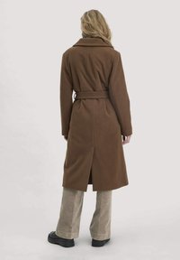 Brauner Wollmischmantel mit tailliertem Gürtel, langen Ärmeln, Rückenschlitz und aufgeschlagenem Kragen, kombiniert mit hellen Cordhose und schwarzen Stiefeln.