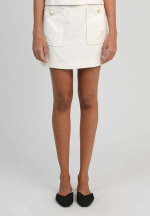 Femme portant une mini-jupe blanche avec de grandes poches avant et des boutons, associée à des chaussures noires à bout pointu et sans lacets.