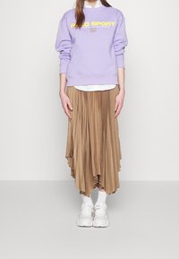 Lila sweatshirt med gul "Polo Sport"-text, matchad med en plisserad, tan-färgad asymmetrisk kjol. Vita sneakers och strumpor fulländar outfiten.