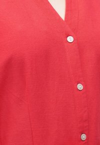 Κόκκινο μπλουζάκι με V λαιμόκοψη και λευκά κουμπιά σε κατακόρυφη σειρά, που δείχνει την υφή του υφάσματος και τις λεπτομέρειες της ραφής.