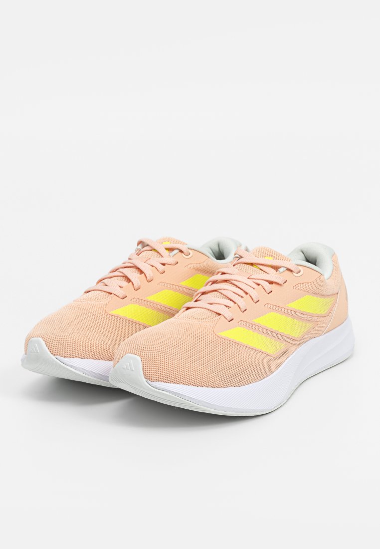 Par de zapatillas de running de malla en color melocotón claro con rayas amarillas, suelas acolchadas blancas y cordones en color melocotón sobre un fondo blanco.
