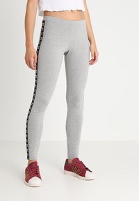 Ben klädda i grå Adidas leggings med svarta logosträck och röda Adidas sneakers med vita sulor, stående framför en vit bakgrund.