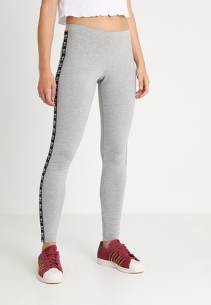 Nohy v šedých legínách Adidas s černými pruhy loga a červených teniskách Adidas s bílými podrážkami, stojící na bílém pozadí.