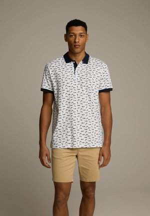 Biała koszulka polo z granatowym kołnierzykiem, wzorzona małymi czarnymi sylwetkami. Połączona z beżowymi szortami, które prezentują luźny krój i swobodny styl.