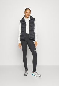 Μαύρο φουσκωτό γιλέκο πάνω από λευκό μπλουζάκι με μακριά μανίκια, συνδυασμένο με μαύρες κολάν και πολύχρωπα αθλητικά παπούτσια. Ορατό λογότυπο στο γιλέκο και τα κολάν.