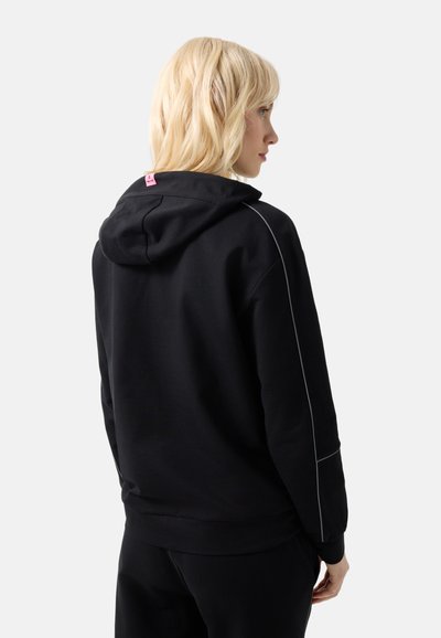 Lyshåret kvinde iført en sort hættetrøje med en lille pink mærkat på hætten, set fra ryggen og siden mod en ensartet lys baggrund.