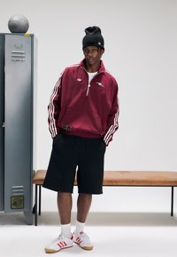 Casaco de malha castanho-avermelhado com riscas brancas, calções pretos e sapatos brancos com detalhes vermelhos. Gorro preto, a pé perto de um cacifo.