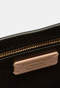 Portafoglio in pelle nera con finitura texture, caratterizzato da una zip dorata e un’etichetta in pelle beige chiaro impreziosita dalla scritta "Coccinelle Authentic Italian Leather."