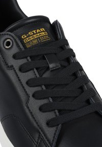 Black leather sneaker featuring flat black laces, subtle stitching details, and a gold branded tag labeled "G-STAR ORIGINALS RAW DENIM."