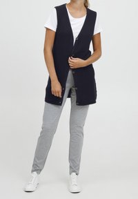 Marineblauer Strickpullover mit Knopfleiste, kombiniert mit grauen Jogginghosen und weißen Turnschuhen. Verfügt über ein strukturiertes Gewebe und hat keine Ärmel.