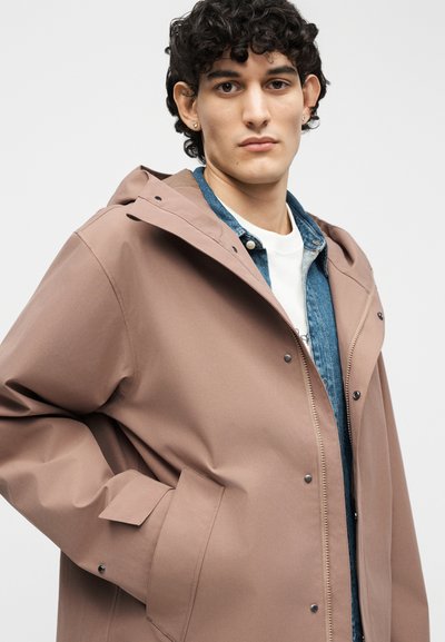 Giovane uomo con capelli ricci che indossa un cappotto con cappuccio marrone sopra una giacca di denim blu e una camicia bianca, che posa con le mani nelle tasche.