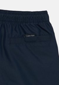 Shorts bleu marine en tissu lisse, présentant une taille élastique, une poche arrière avec une étiquette noire sur laquelle est inscrit "Calvin Klein".