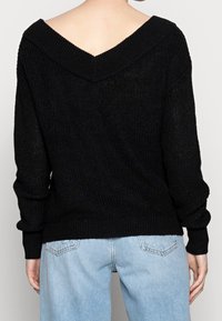 Pull noir en tricot avec un large col en V et des manches longues, doté de poignets et d'un ourlet côtelés. Porté avec un pantalon en denim bleu clair.