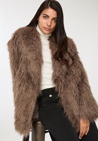 Cappotto in finto pelliccia marrone con una lunga e shaggy texture; indossato sopra un dolcevita bianco, abbinato a jeans neri. Presenta un design con apertura frontale.