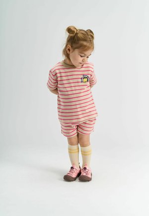 Pasi s kratkimi hlačami in majico v bež in roza črtah, ki ima okroglo ovratnik in potisk; v kombinaciji z kremnimi nogavicami do kolen ter roza čevlji.