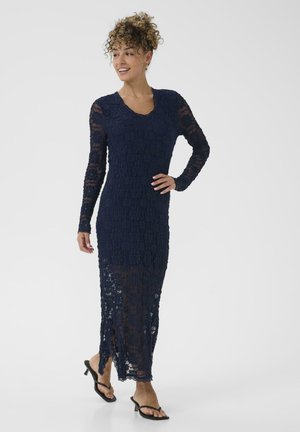 Robe en dentelle bleu marine longue avec des manches longues, présentant un décolleté arrondi, une texture transparente et une fente sur le côté, associée à des sandales à talons noirs.