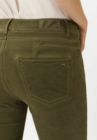 Pantalones verde olivo con una textura suave, con un bolsillo trasero y un detalle de cinturilla acentuado con metal. Las costuras son sutiles y refinadas.