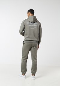 Άνδρας με την πλάτη γυρισμένη, φοράει πράσινο ελιάς φούτερ με κουκούλα και ταιριαστά παντελόνια φόρμας, με λευκά αθλητικά παπούτσια σε λευκό φόντο.