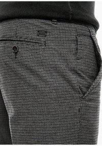 Detailní záběr na kalhoty s šedým a černým houndstooth vzorem, s bočními a zadními kapsami, černým knoflíkem a malou černou značkovou štítkou na pase.